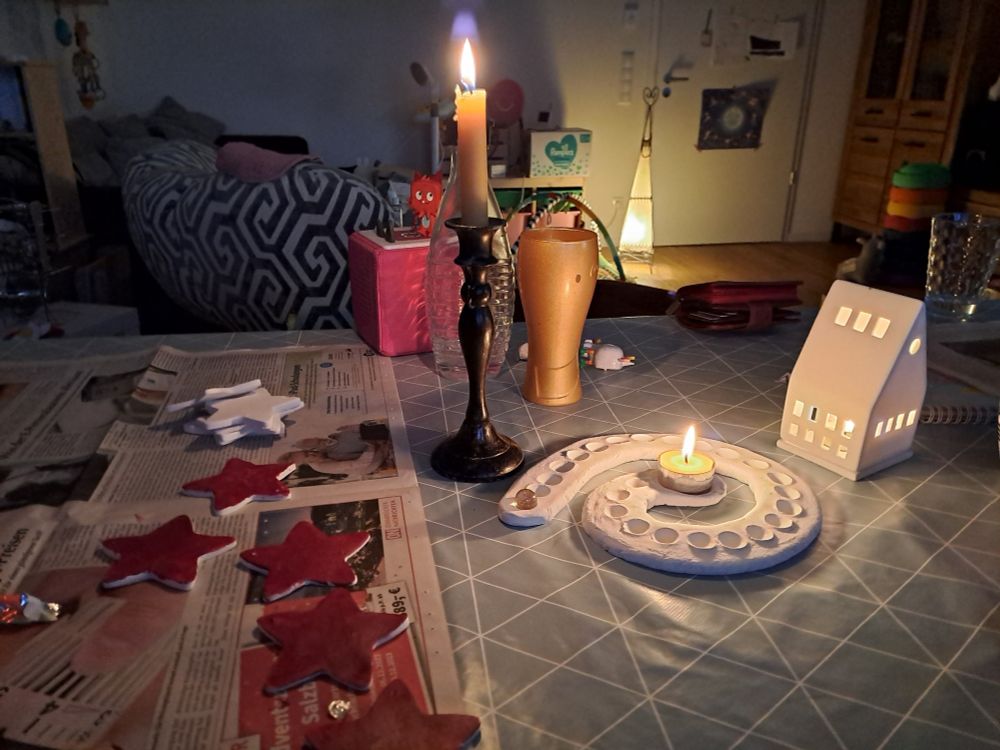 Ein Tisch, auf dem drei Kerzen brennen, eine in einem schwarzen Kerzenständer, ein Teelicht in einer Adventsspirale und eines in einem kleinen weißen Häuschen. Daneben ein paar teils rot angemalte Sterne aus Modelliermasse auf einer Zeitung. Dahinter eine pinke Toniebox, auf der eine Toniefigur steht, eine weitere liegt daneben. Außerdem sind da ein goldenes Glas und eine Flasche. Im Hintergrund ist ein Wohnzimmer zu erkennen, in dem eine gedämpft leuchtende Lampe auf dem Boden steht.