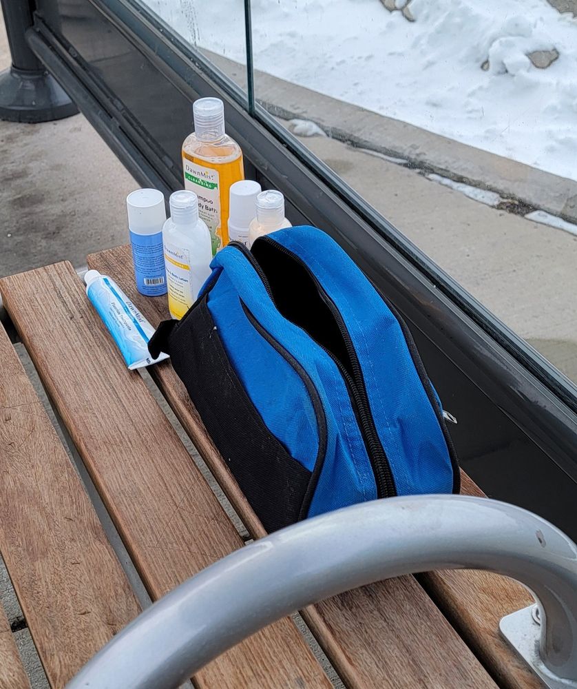 New toiletries, likely freebies from a hospital stay given the institutional branding, are arranged on a wooden bench at the Golf Mill bus station in Niles, Illinois. An empty blue toiletries bag is open next to the hygiene items.