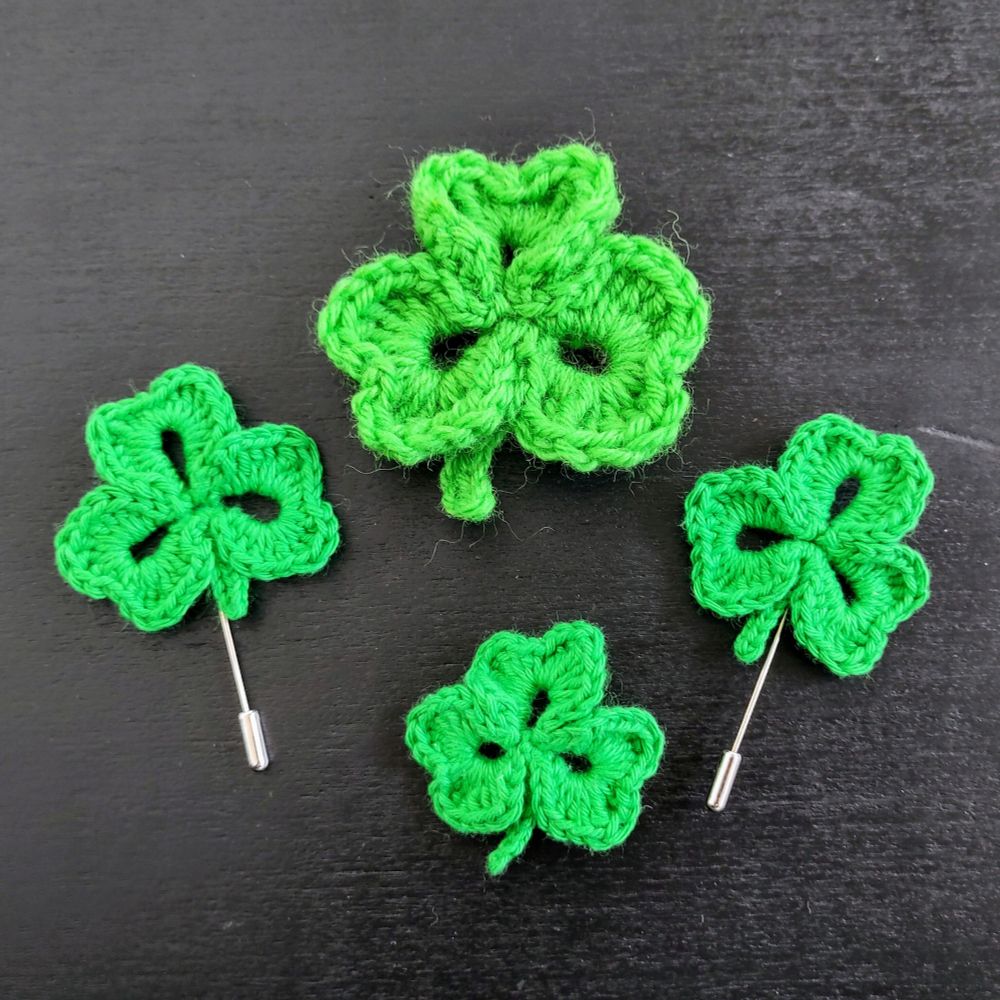 Four green crocheted shamrocks, one large, three small, two with badge pins visible 
