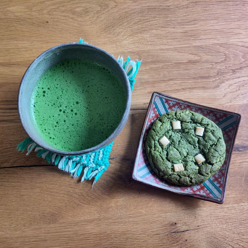 Matcha tea in a bowl next to a small plate containing a matcha and white chocolate cookie