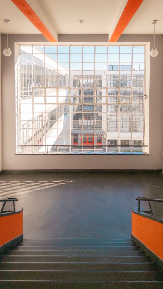 View of a large, grid-patterned window overlooking Bauhaus entrance, seen from the top of a stairway with orange railing accents.
