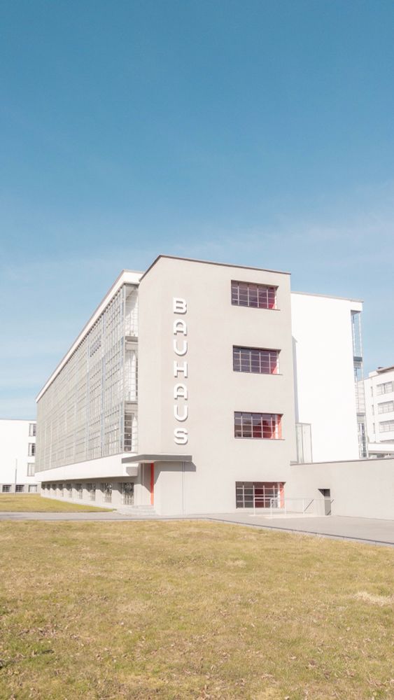 Bauhaus building with a modernist architectural style, featuring a gray facade, grid windows, and a large sign reading "Bauhaus" in vertical text. Grass lawn in the foreground.