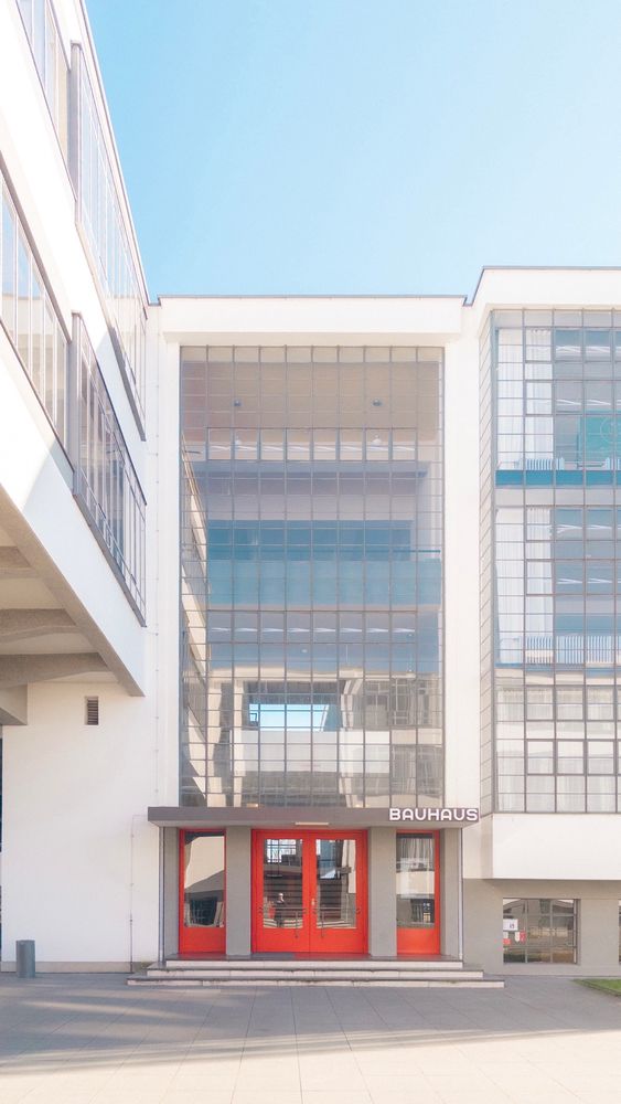 Exterior of the Bauhaus building in Dessau, featuring large glass windows, a beige facade, and red double doors under the Bauhaus sign.