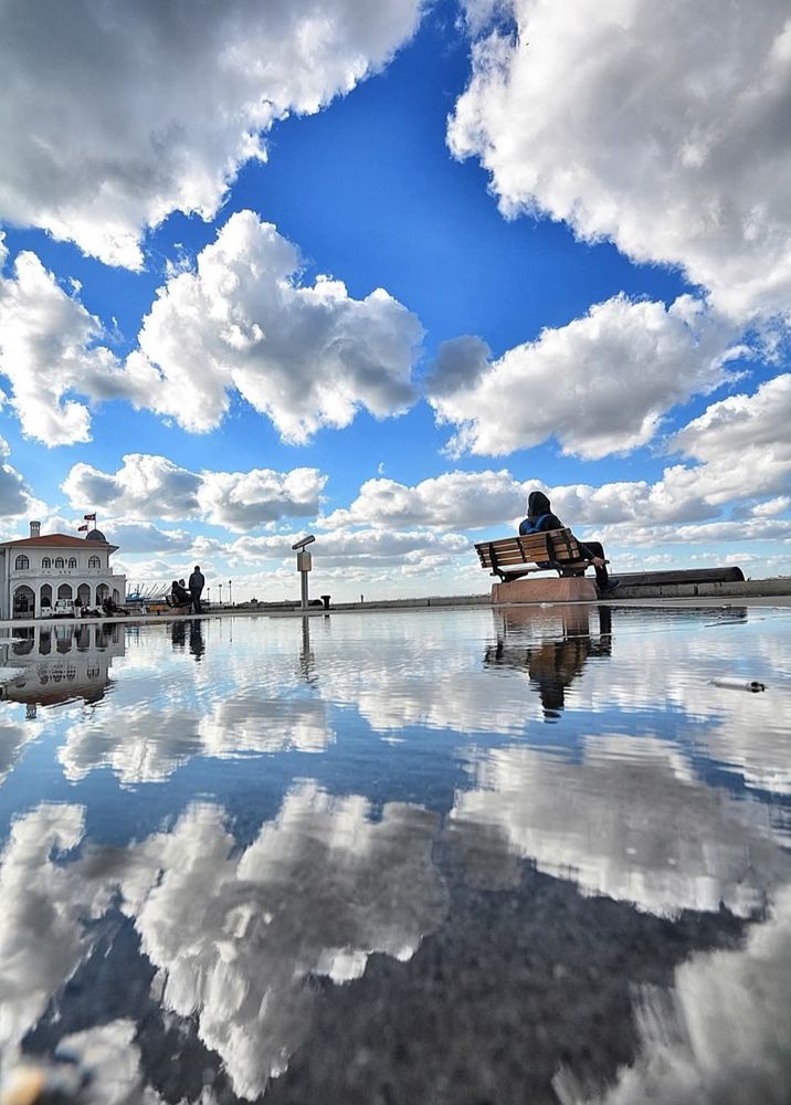 An urban photo with a person sitting on a bench with bright puffy clouds that are reflecting on pavement, in water, after it rained in Istanbul, Turkey.