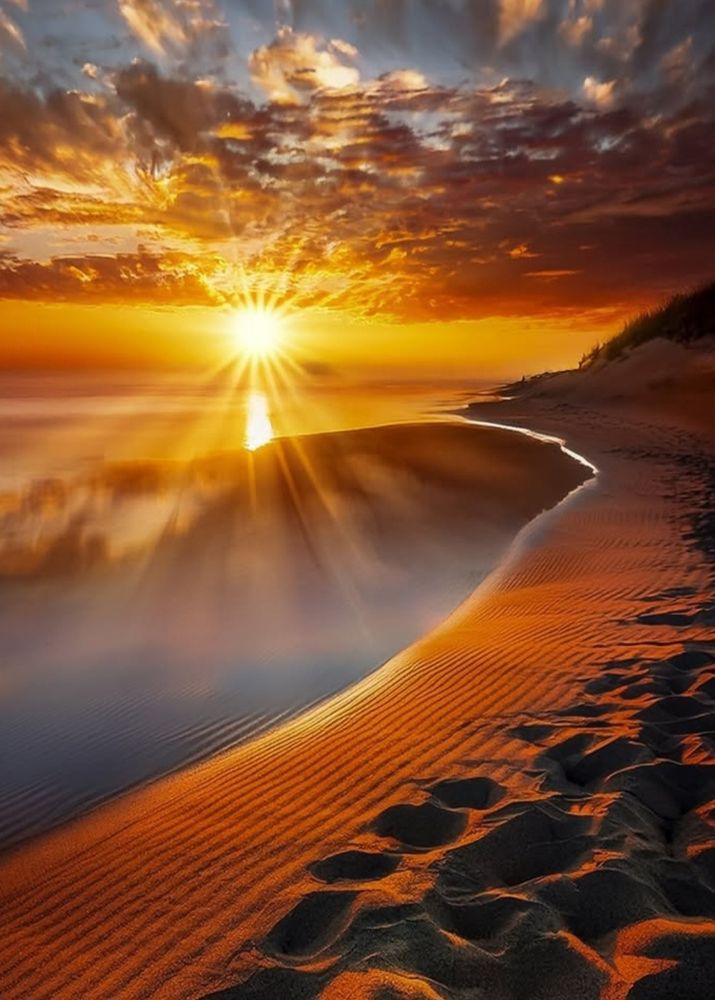 A photo of a beach with a brilliant sun, calm water & beautiful orange clouds.