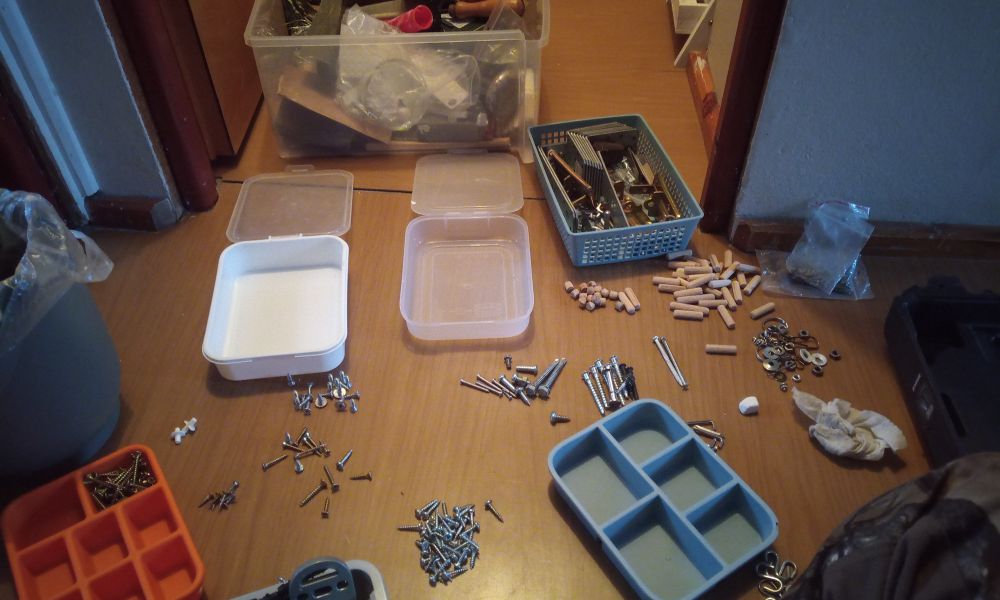 Several plastic boxes with compartments for screws and other little things, wooden bits with said material piled up and scattered around me. I just went through two tiny organizers from IKEA, orange and blue-gray one and tossed damaged pieces. There is a shallow basket of the same blue-gray colour with heavy duty pieces of metal squares and L brackets for connecting and supporting wooden structures, simple small door latches I use for windows in stormy weather. On the left there is a compost bin in the shape of an urn, I cannot help it and unsee the similarity, it's of a green-gray lichen colour.