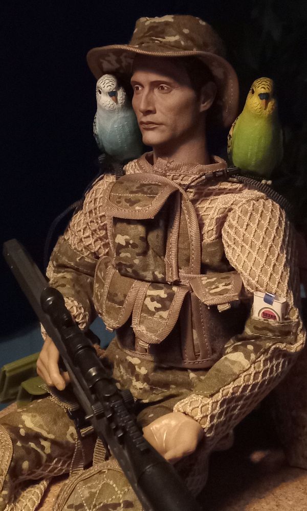 Action figurine with a custom head sculpt and took a garderobe from an unsuspecting soldier toy to give my Mads some basic layer, tactical vest, box of cigarettes, sniper rifle, hat and.. two budgies, blue and green, perched on his shoulders. He also sits under a lamp that emits warm light.