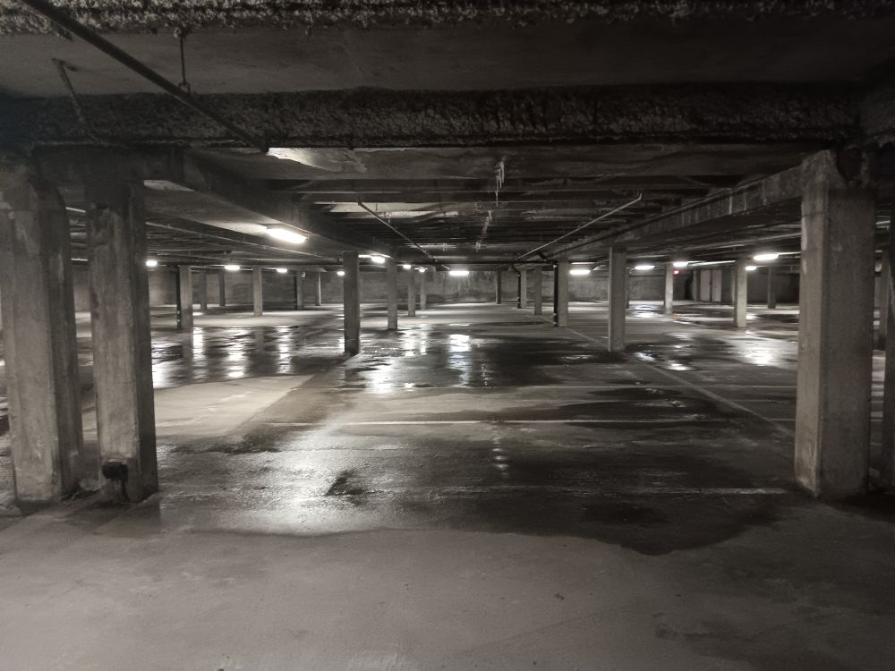 A parking garage with a low roof, fluorescent lighting, and numerous puddles from the leaky ceiling. There are absolutely no cars or people.