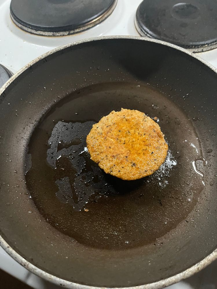 Auf einem weißen Herd mit vier Platten steht eine dunkelbeschichtete Bratpfanne, darin fett und ein rötlicher Klops