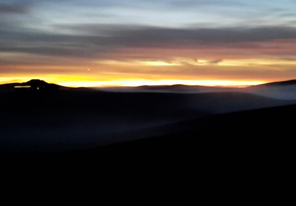 This photo is taken on the rolling hills in southeast Washington state. It is on a upper hill and you can see the fog set down in the valleys with the tops of the hills poking up. Bright orange, Yellow horizon line with thin clouds above it glowing pink slowly fading into gray against a dusty gray-blue sky. 