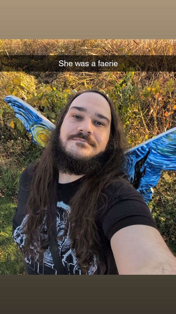 A long haired, beaded man wearing all black. He's taking a selfie in front of some faerie wings