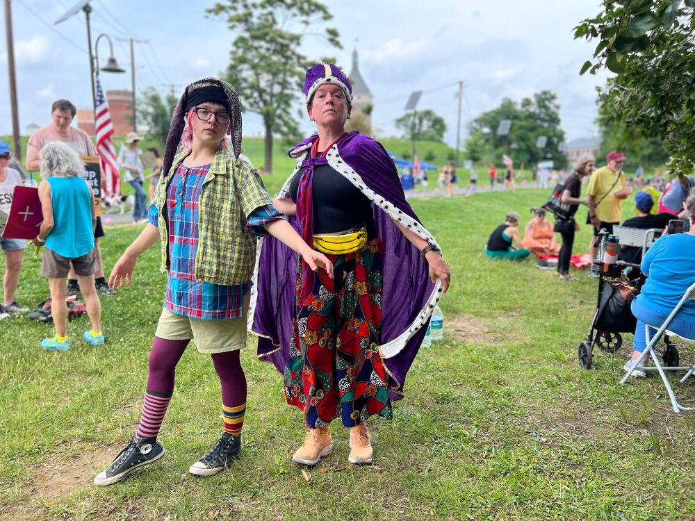 A young person in jester attire and a woman in a people king robe and crown at Ft Reno (DC) No Kings community picnic