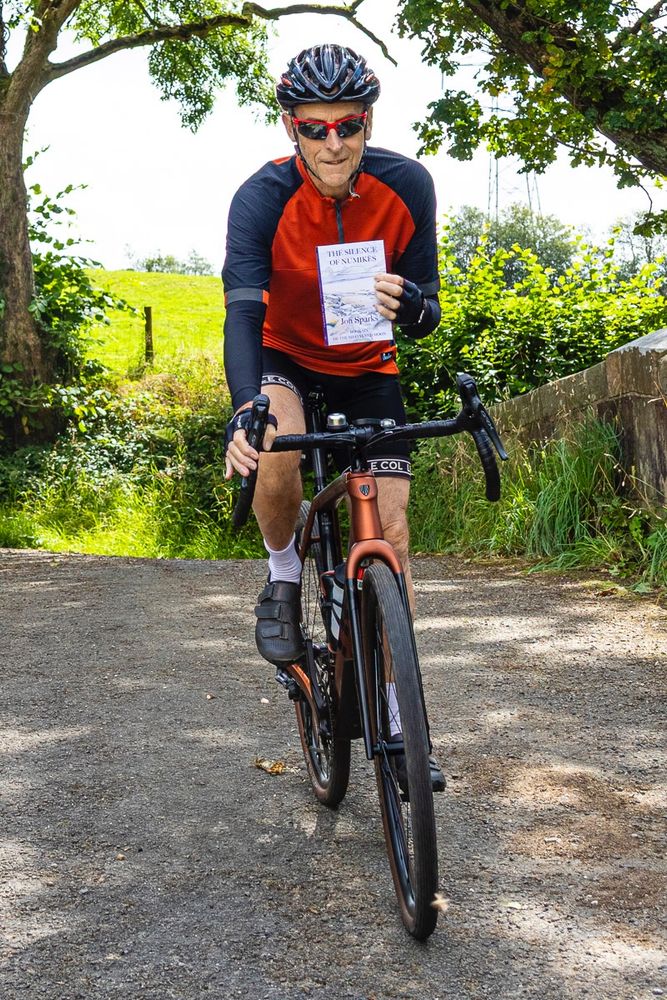 Photo shows author in orange and black cycling jersey riding over a stone bridge with a backdrop of field and trees, holding a copy of The Silence of Numikès in his left hand.

