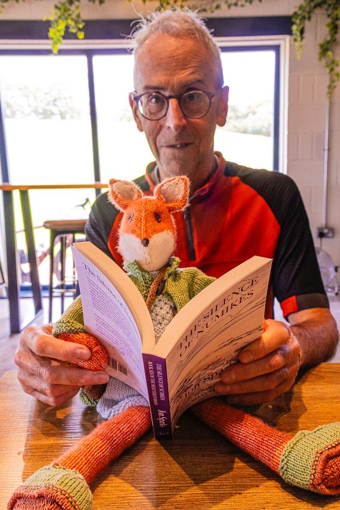 Photo shows author in orange and black cycling jersey holding soft toy fox in white dress and green cardigan. The fox is reading The Silence of Numikès (with a little help from the author).