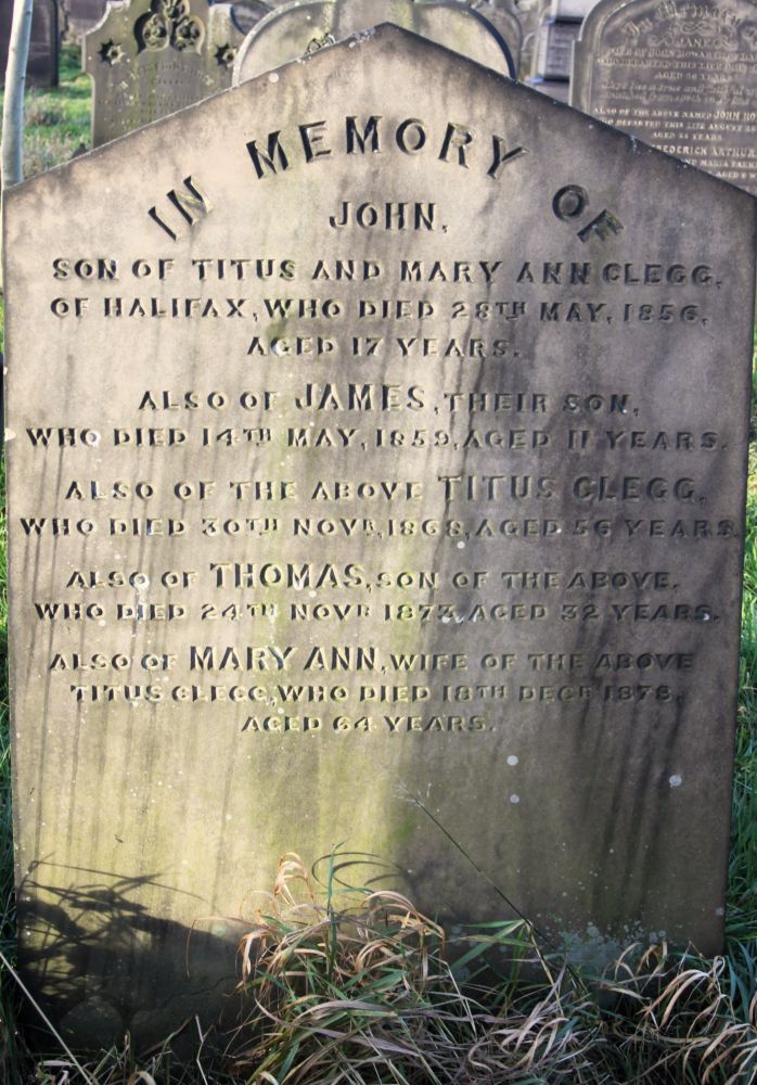 The image shows a close-up of a weathered headstone at Lister Lane Cemetery which marks the burial place of members of the Clegg family. The monumental inscription reads: "In memory of John, son of Titus and Mary Ann Clegg. of Halifax, who died 28th May 1856, aged 17 years. Also of James, their son, who died 14th May 1859, aged 11 years.  Also of the above Titus Clegg, who died 30th November 1868, aged 56 years. Also of Thomas, son of the above, who died 24th November 1873, aged 32 years. Also of Mary Ann, wife of the above Titus Clegg, who died 18th December 1878, aged 64 years."