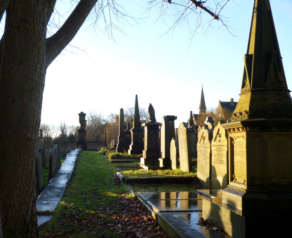 An image taken at Lister Lane Cemetery in November 2024. It shows a view along the central grassed path leading towards a pair of large stone monuments which flank either side of the steps leading down to the cemetery's lower section. Stone headstones and grave monuments bathed in autumn sunlight line the path to the right. On the left in the foreground stands a tree trunk, beyond which other headstones can be seen. 