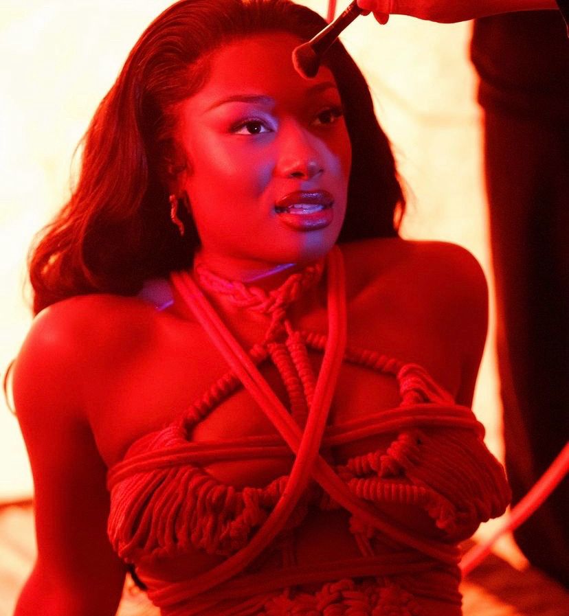 bts image of megan in a similar red string outfit getting makeup put on her face. she’s illuminated by red light and she’s so beautiful