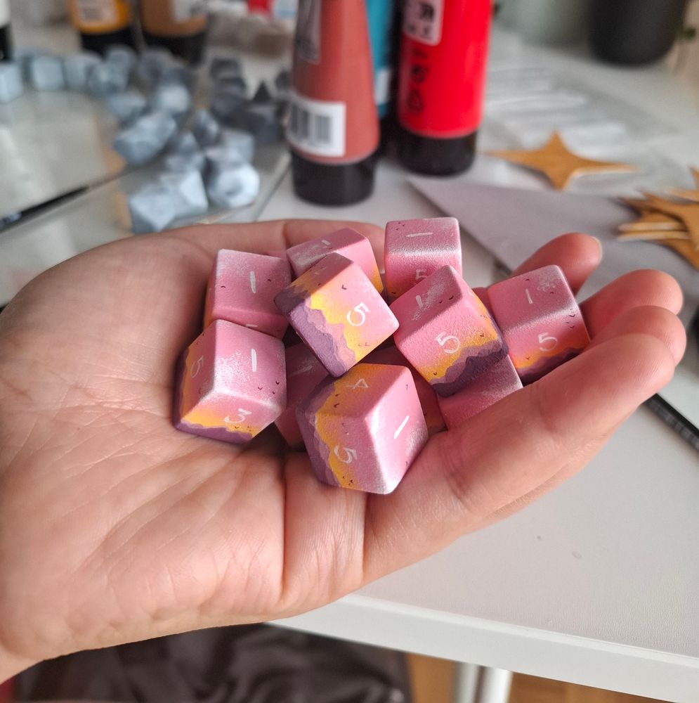 A hand holding a pile of d6. They are painted to look like a mountain range in front of a sunset. A few tiny birds are flying in the sky