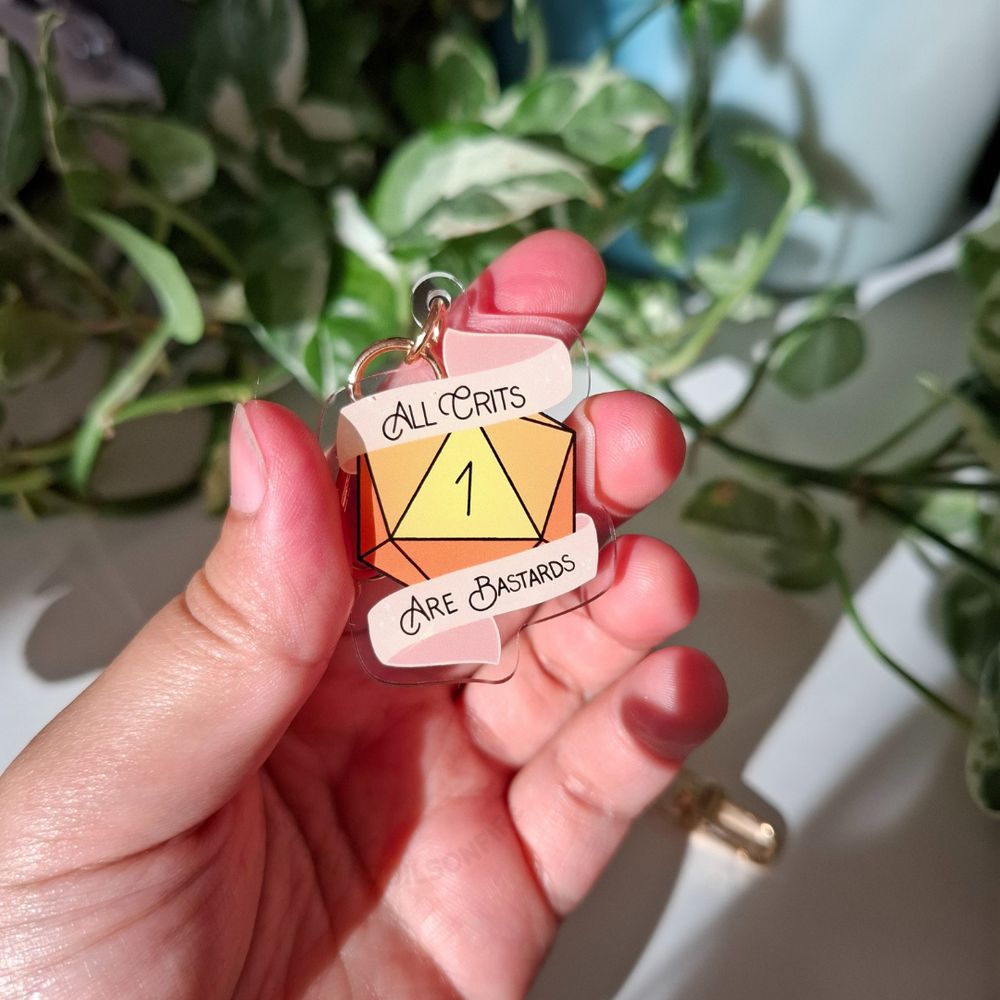 a hand holding a keychain. it's a d20 showing a 1. a banner says "all crits are bastards"