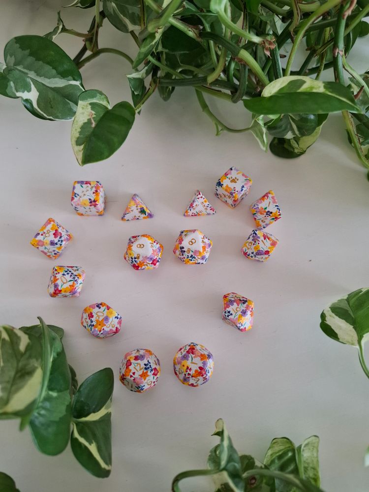 Two polyhedral dice sets arranged to form a heart. They are hand-painted. The base is white and colourful flowers run along the edges. The numbers are painted in gold. Instead of the number 20 it shows interlocked wedding rings. 