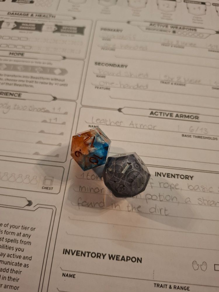 Two d12s on a daggerheart character sheet. One of them is orange and blue, with white clouds inside. The other one is clear, with a moon inside
