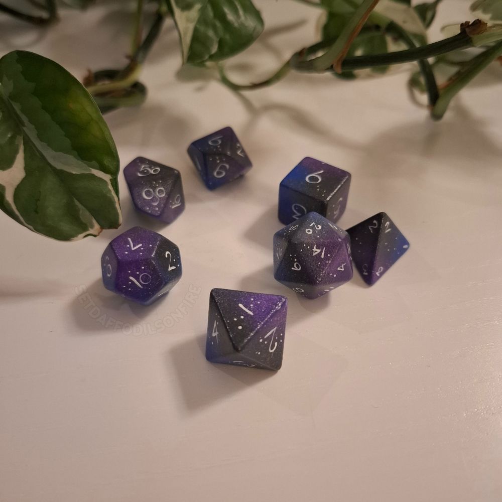 a handpainted polyhedral dice set. It's painted in a galaxy style with purple, blue and dark grey colours. the numbers are white and there are starry patterns across it.