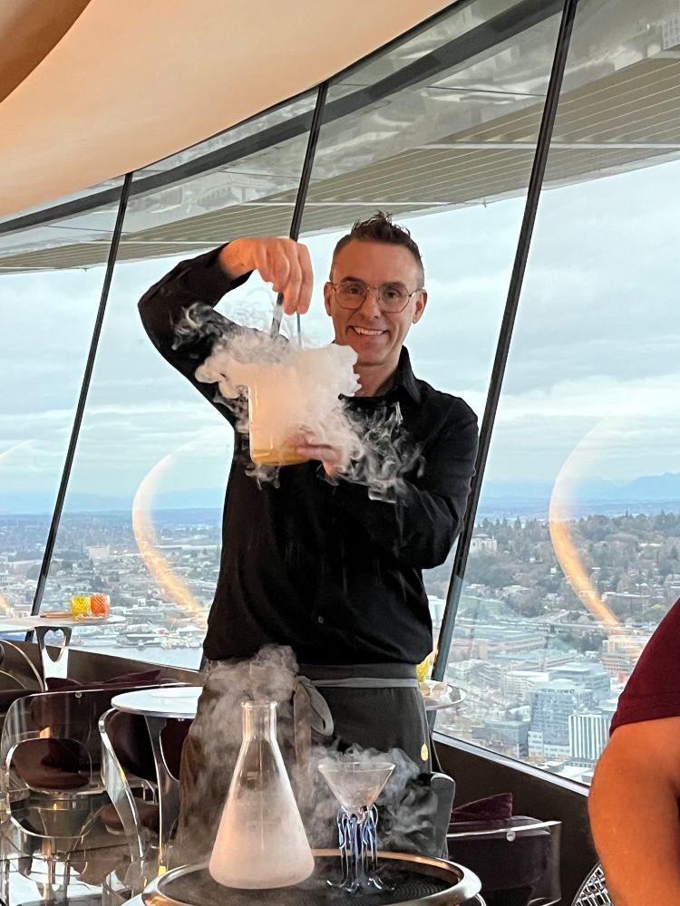 Our waiter mixing some fancy drink. A beaker is on a tray in front of him nex to a martini glass with octopus legs, smoke is coming out of the beaker. The waiter is holding a big glass with smoke coming out & he is stirring the liquids inside