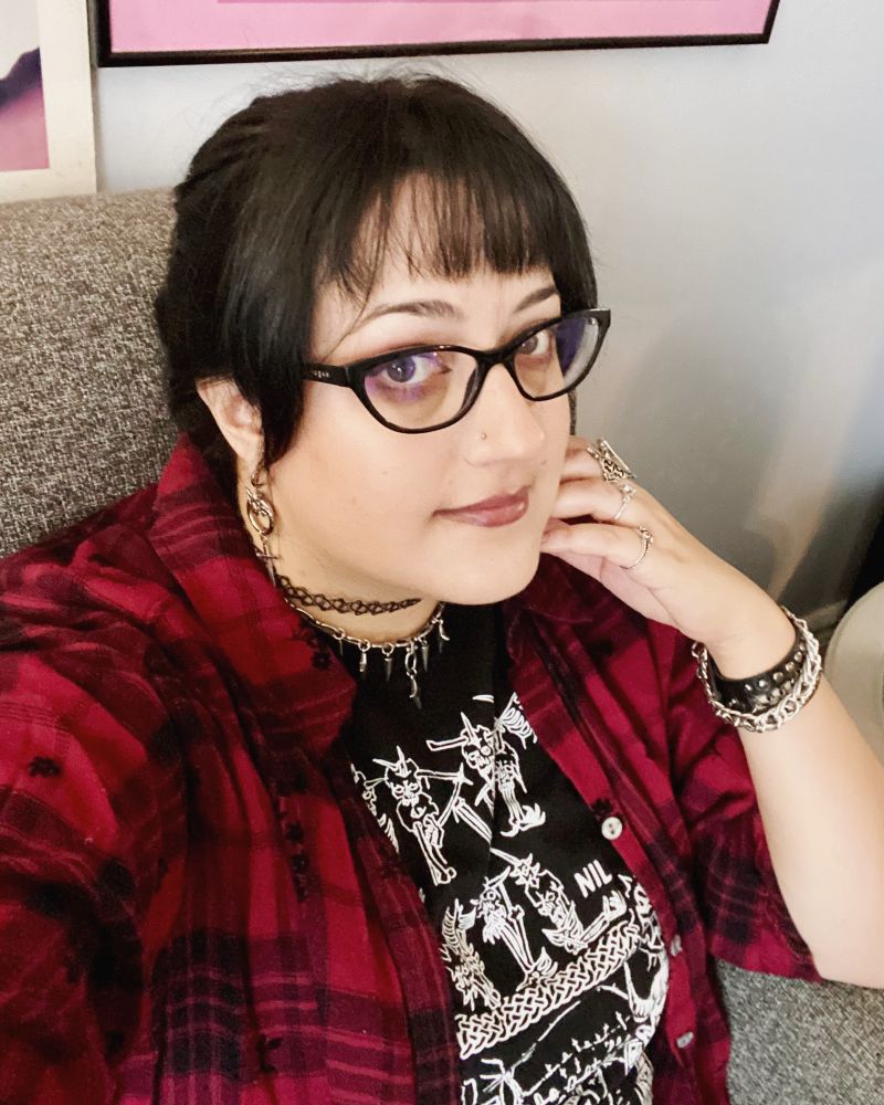 A selfie of a pale skinned white woman with black hair in a ponytail and a messy fringe and thick rimmed glasses sitting on a couch. She has a red plaid shirt on, a black band t shirt and several black and silver jewelry pieces