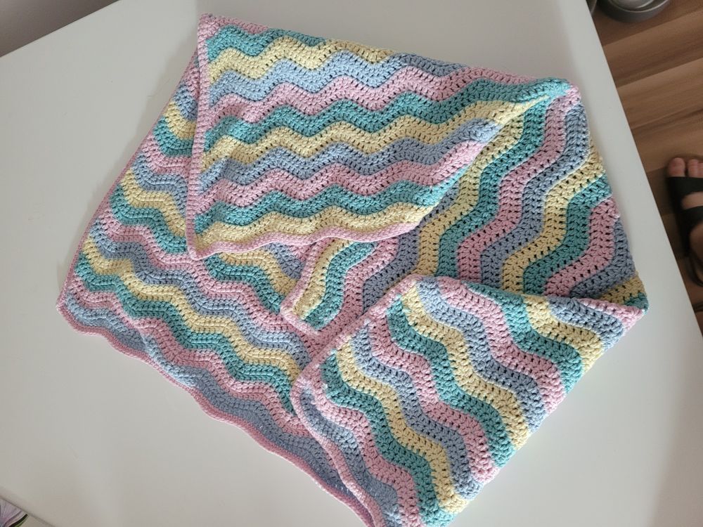 Gehäkelte Babydecke im Wellenmuster mit anwechselnden Farben blau, rosa, grün und gelb. Drumherum ist ein Rand in rosa gehäkelt.