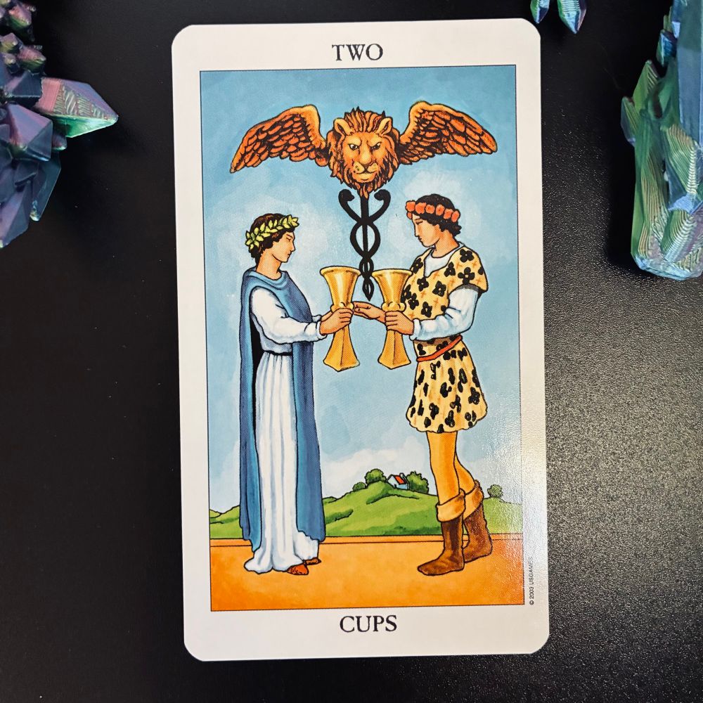 A tarot card. The 2 of cups. A man and woman each holding a large golden cup. They seem to be in the midst of exchanging them.