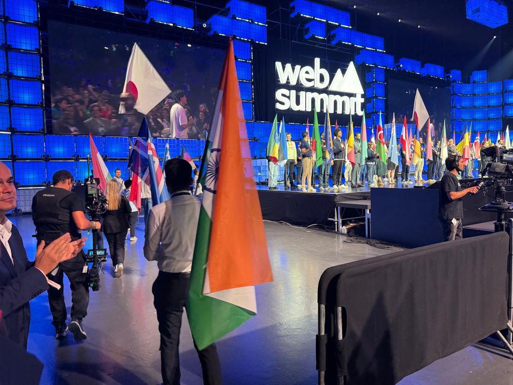 People holding various national flags, including India’s, are walking onto the Web Summit stage as camera operators film the procession. The blue “web summit” logo and numerous flags are clearly visible in the background.