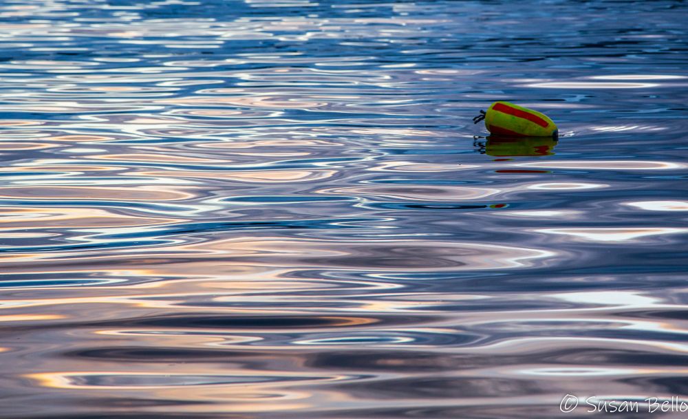 A single yellow and red buoy bobs in a rippled sea that goes from blue to light orange in the light of sunset. 