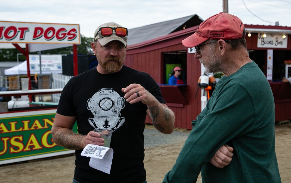 A man in a black shirt with a white image of a diving helmet with a ball cap and sun glasses on his head talks to a man in a green shirt with orange hat. Behind them is a booth selling hot dogs and a red booth.