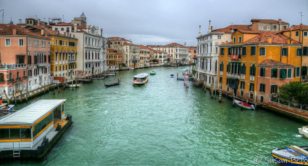 Green water framed by orange, white, and pink buildings on the sides of the canal beneath a gray sky. Several boats are traversing the canal.