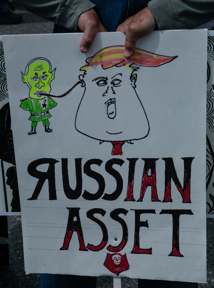 Hands hold a sign with caricatures of Trump and Putin with Putin's tongue connected to Trump's ear. The text reads 