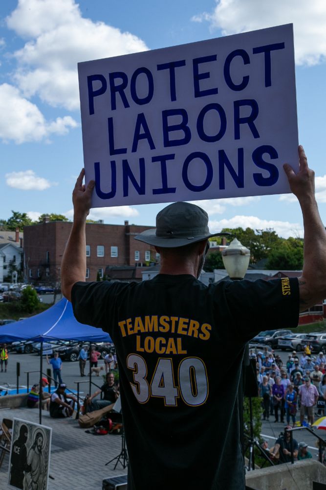 A man in a Teamsters local 340 shirt stands on top of a hill in front of a crowd holding a sign that reads 
