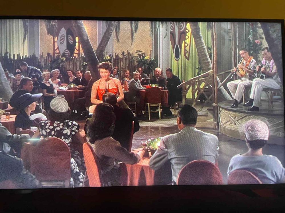 Actress May Wynn plays a sultry nightclub singer in a red gown, performing in a tiki lounge decorated with palm trees and tribal masks, accompanied by musicians wearing Hawaiian shirts and leis, in a scene from THE CAINE MUTINY