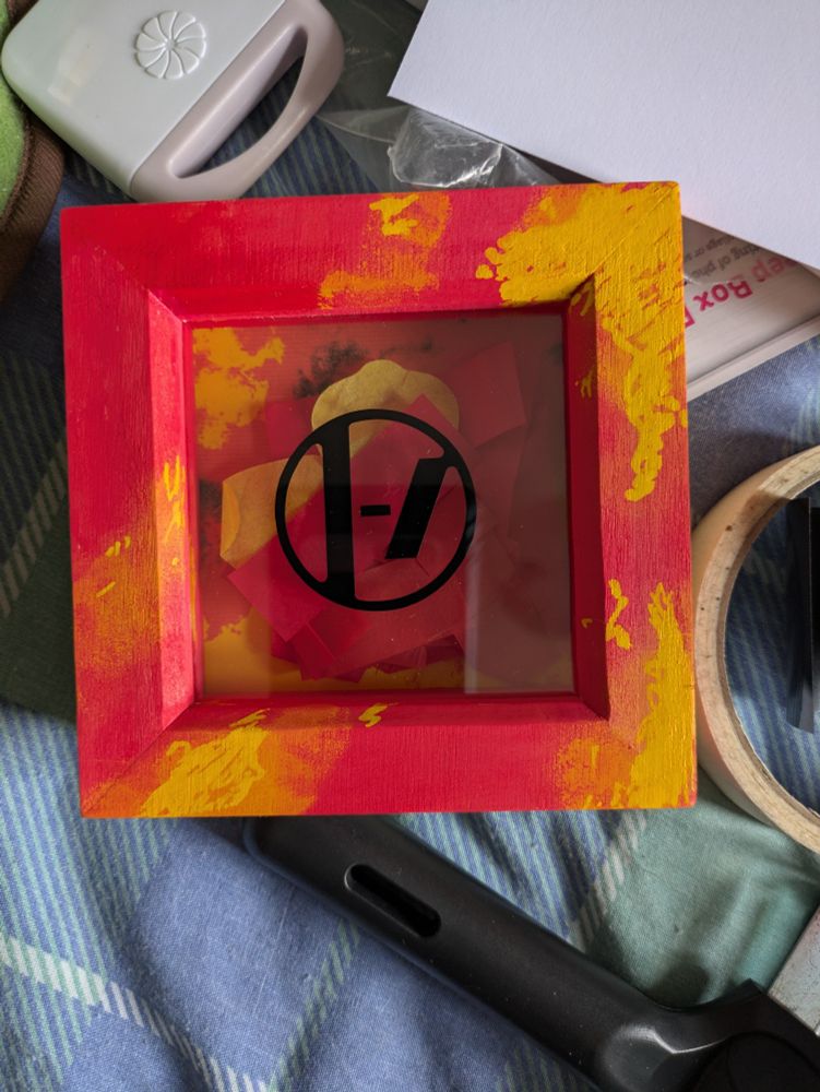 A 9x9 cm shadow box painted red and yellow to match the Clancy album cover. The twenty one pilots logo is in the middle of the glass. Inside it is red and yellow confetti. The box is on a blue and green duvet, surrounded by scattered art supplies.