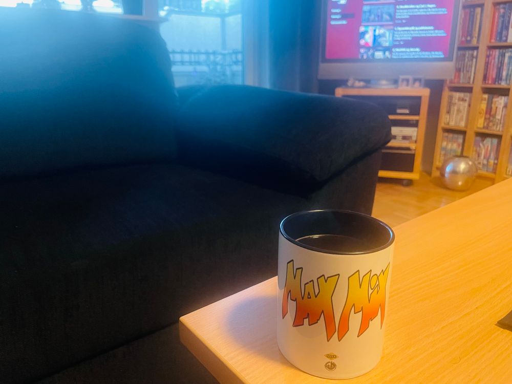 Kaffekopp med teksten Max Mix, foran en del av en sofa og tv-skjerm.