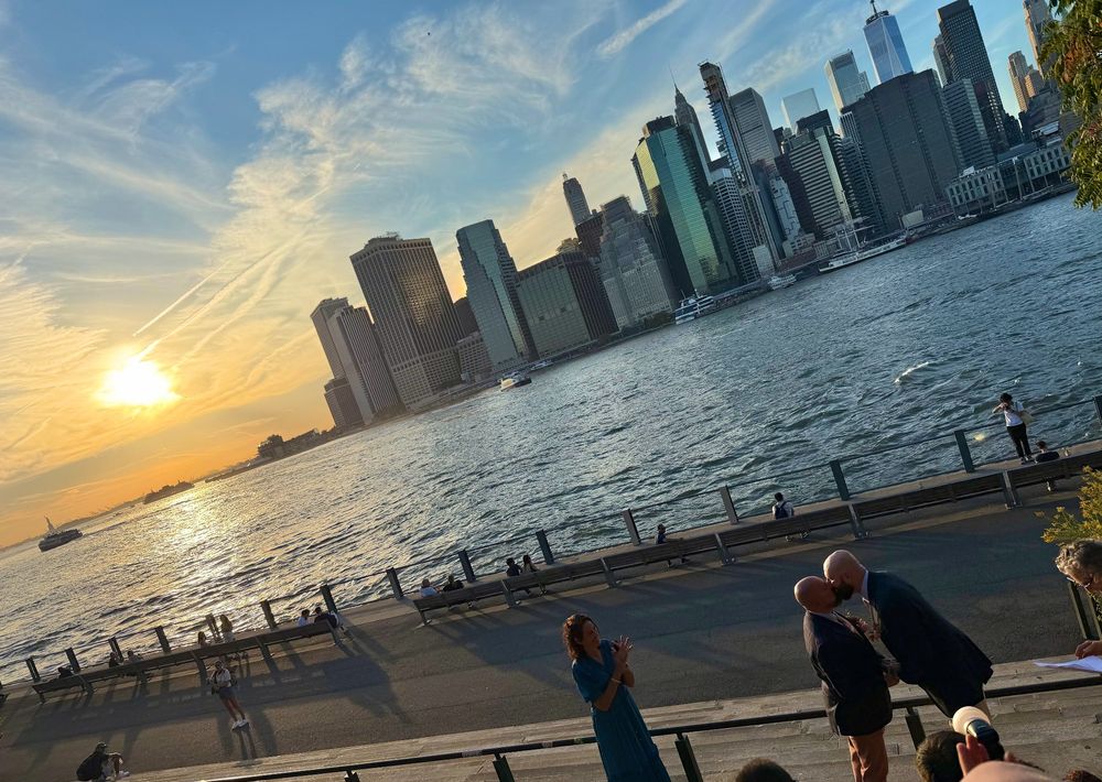 two men getting married with the manhattan skyline and the statue of liberty behind them