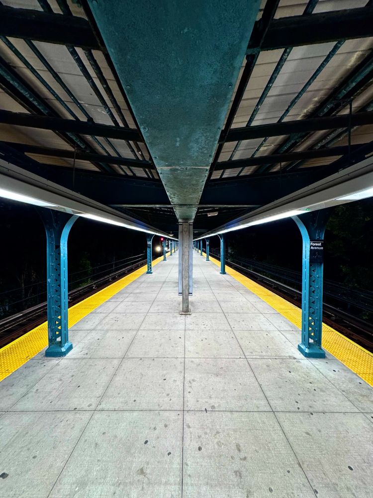 very symmetrical picture of the platform at Forest Avenue station in Queens. A subway train is approaching in the distance. 