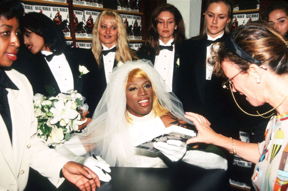NBA player Dennis Rodman wearing a wedding dress and blonde wig, rocking some elbow length white gloves, seated and doing a press spot about his marriage to himself. He has a wedding party of attractive women around him, all are wearing tuxedos. A woman in the foreground is I believe a clerk accepting signed documents, she is absolutely of a time with a glasses chain, dress, and headband that scream “I am a 90s librarian”. 