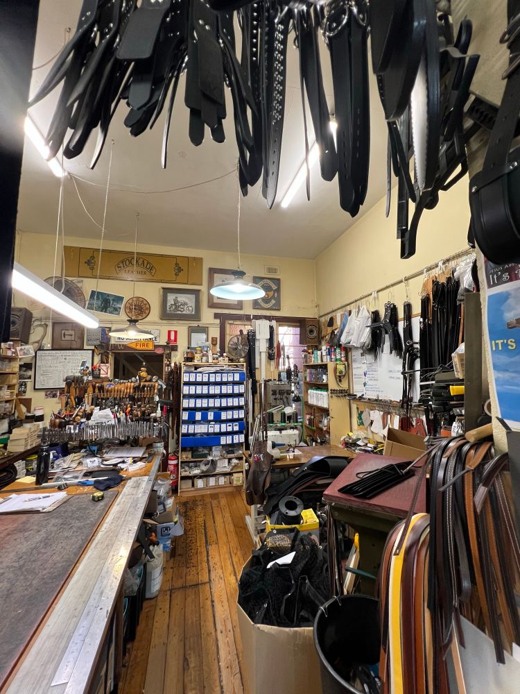 Interior workshop view showing belts hanging from the ceiling, a long workbench cluttered with leather tools and papers, shelves stacked with supplies, and a central aisle leading toward the back of the shop.