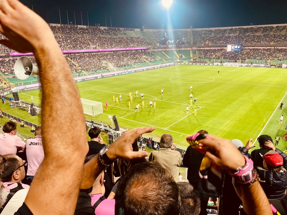 Arms of fans aloft in the Curva Nord Superiore, as the Palermo players come close to a goal on the pitch below, under the floodlights of the Stadio Renzo Barbera in Palermo 