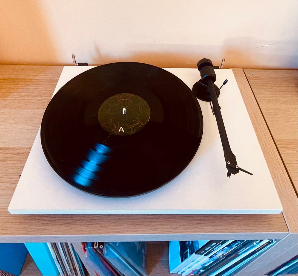 Side A of an LP copy of the Liverpool band The Shipbuilders 2nd album This Blue Earth sits on a white turntable on shelves with more LPs stored below 