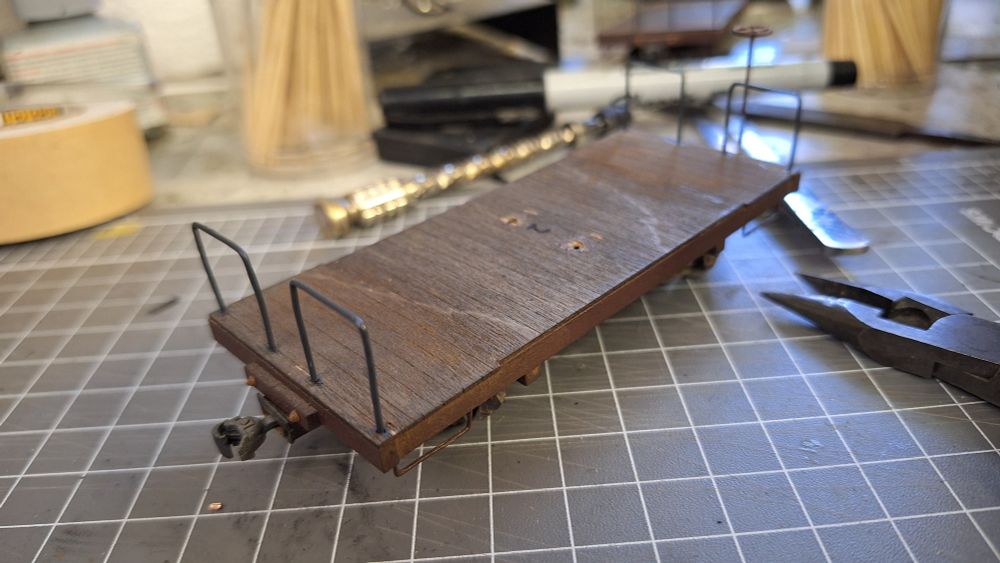 A model caboose chassis sits on a workbench, with four handrails bent out of copper wire painted with dark grey enamel.