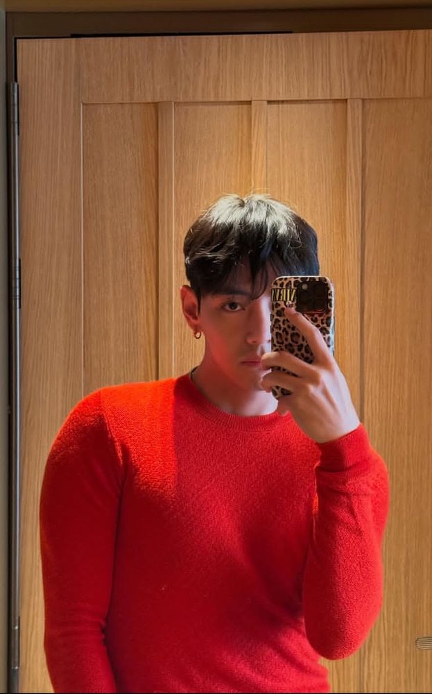 A muscular Taehyung wearing a very tight red long sleeved shirt taking a mirror selca. One earring dangles from his ear. His phone case is leopard print.