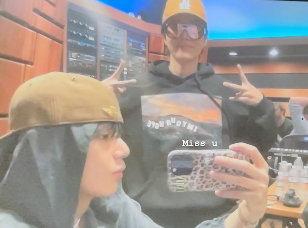 A grainy, cloudy picture of Taehyung and Hobi in recording studio. Tae is holding is phone up to take the pic. He's wearing a hoodie with the hood up and a backwards ball cap over that. Hobi is wearing his hat properly and flashing two peace signs. Text added to the picture reads: Miss u