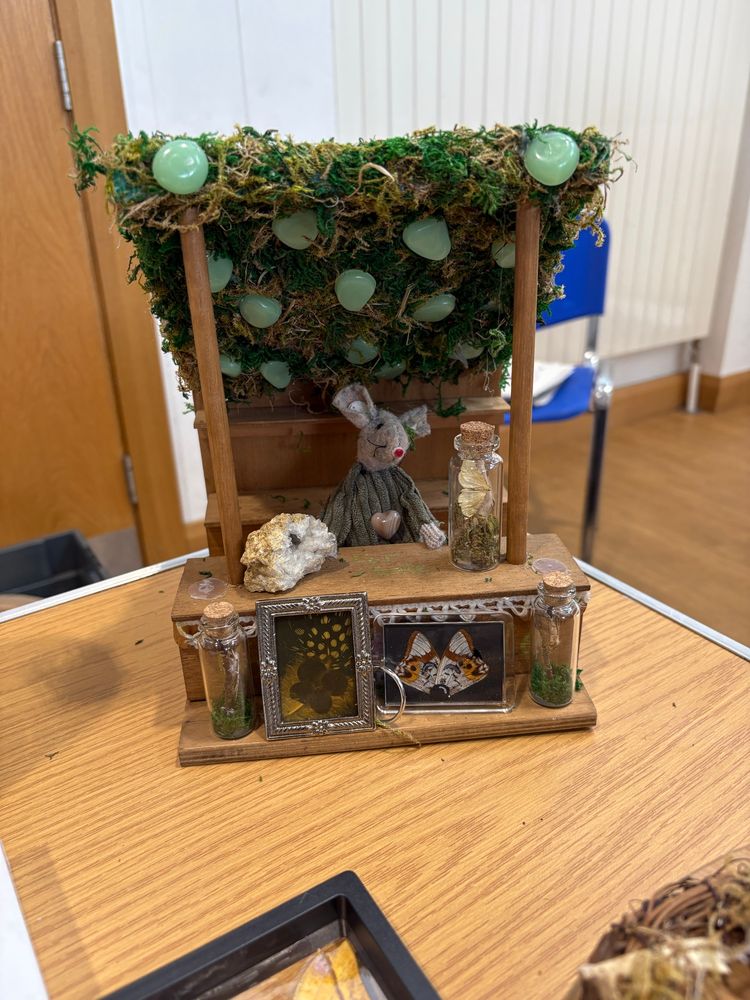 Tiny market stall made from wood & moss, with green crystals studded along the roof. A tiny felt mouse model in a green jumper is selling various crystals, butterflies in jars and framed butterflies. 