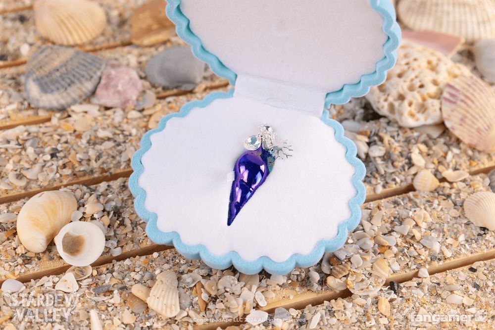 A photo of the Mermaid's Pendant in its pale blue and white clamshell keepsake box. It's an iridescent plated glass pendant on a silver-plated chain. The box is resting on small wooden planks strewn with sand and shells.